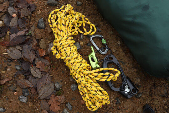 Top view flat lay of professional climbing gear on ground background including coiled yellow nylon rope, metal screw gate carabiner, green belay device and black hand ascender clamp in forest