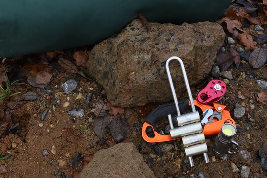 Horizontal view of professional climbing gear on ground near big stone in forest including silver brake bar rack descender, orange hand ascender rope clamp, red pulley block and led flashlight