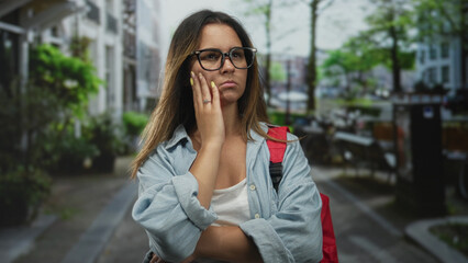 Fototapeta premium Young woman touching cheek with hand, wearing glasses and red backpack on city street in casual denim shirt and white tank; pensive doubt.