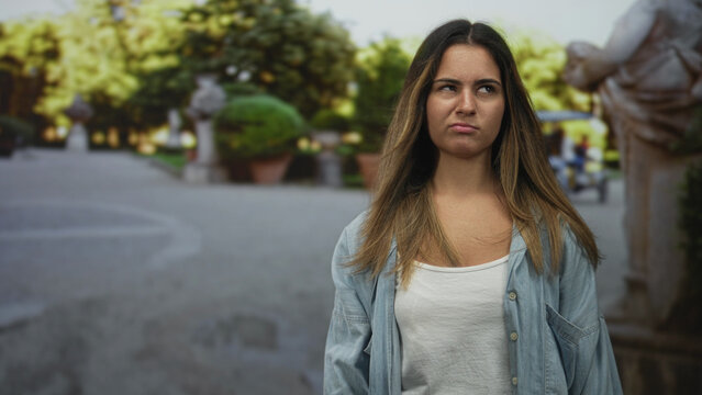 Woman grimacing with eyes squinted, bare upper chest visible in white tank and denim shirt on street lined with trees and stone statues; disgust urban frustration.