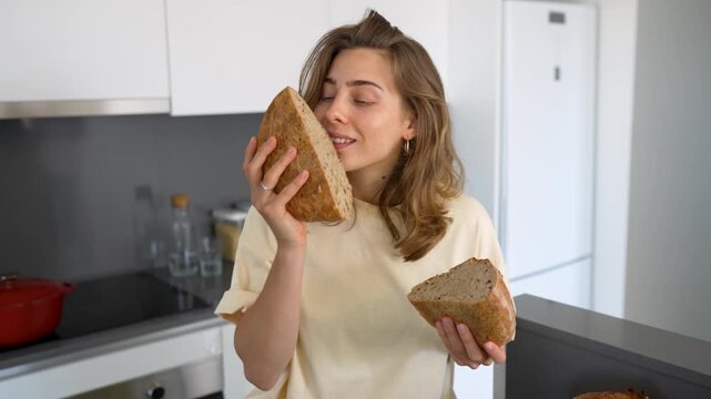 Young woman stands in modern kitchen, cutting freshly baked bread and enjoying its aroma. A housewife has baked sourdough bread in a cozy room. Concepts of cooking, comfort, and preparation.