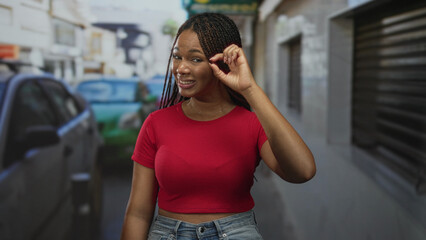 Fototapeta premium Young woman in red shirt pinching braid with fingers on a busy city street among parked cars and storefronts; skeptical confidence.
