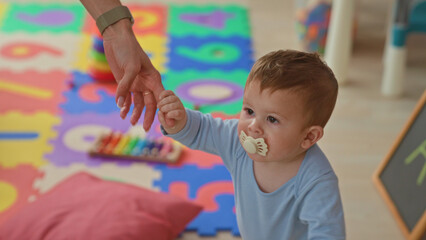 Obraz premium Boy toddler holds adult hand while standing with pacifier in mouth on a colorful foam mat in a building playroom; first steps joy.