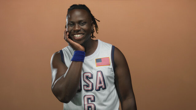 Man touching cheek and smiling in a studio wearing a usa basketball jersey against a peach backdrop; joyful confidence.