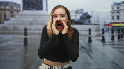 Fototapeta premium Redhead woman calls out with hand to mouth on wet street near building and statue, wearing casual jeans and studded belt; youthful joy.