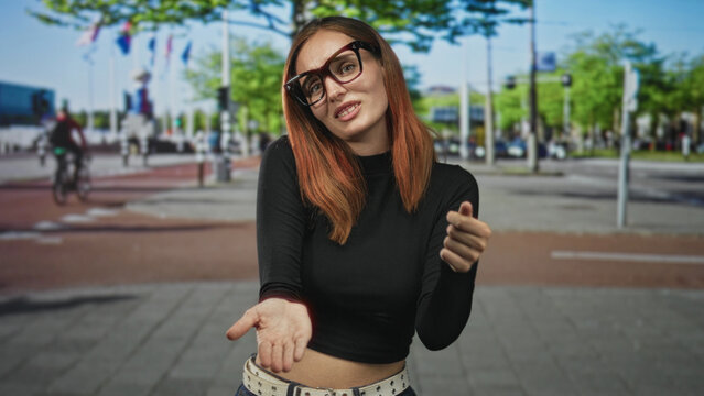 Young redhead woman in glasses touches chest with both hands and offers palm forward gesture on an urban street; apology sincerity.