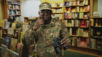 Young black soldier man holding binoculars with finger to eye while standing among crowded bookshelves in bookstore  vigilance duty. © Krakenimages.com