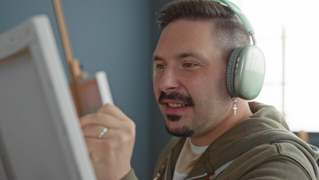 Man paints a canvas with a brush while wearing large headphones and a cross earring in an artist studio; concentration creativity.
