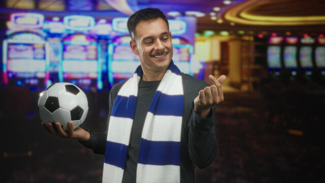 Moustached man holding soccer ball and showing finger heart gesture while smiling and wearing blue white scarf in building; passion betting celebration.
