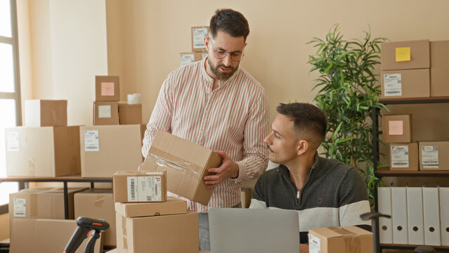 Two men packing boxes, standing man holds a cardboard box while seated man shows phone with hand raised in a building; teamwork productive.