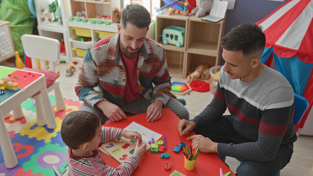 Young boy places wooden puzzle pieces while two men guide with hands at a colorful play table in a building; warmth bonding.