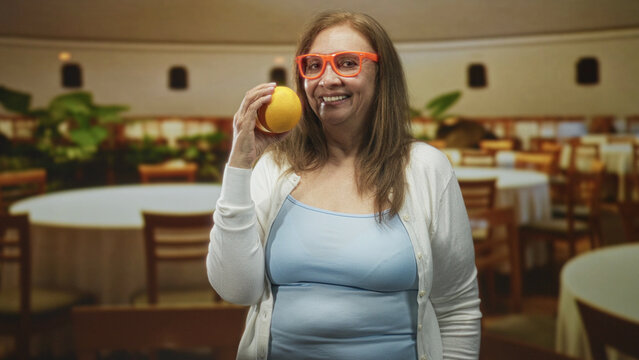 Senior hispanic woman holds orange to cheek with hand visible and smiles in a building banquet room while wearing orange glasses; joy playfulness.