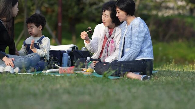 Warm sunlight glows on green grass. Women and a child sit close, sharing food. Laughter and chatter fill the air. They enjoy calm, shared moments outdoors. Ideal for family, leisure, or nature themes