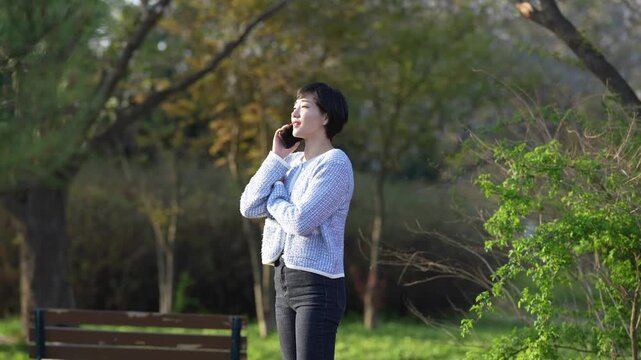 Woman talks on phone near park bench. Trees surround her in green daylight. She stands relaxed, unaware of nearby nature. Soft light enhances peaceful, casual moment