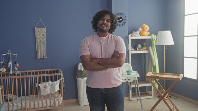 Man in pink t shirt standing with arms crossed by a wooden crib, mobile and toy shelf in a studio baby room with changing table and floor lamp; quiet pride.