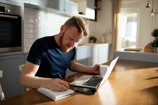 Young man writing notes while working on laptop at kitchen table