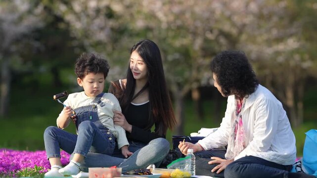 Spring cherry blossoms frame a joyful family picnic. Young child, held gently, gazes at a smartphone. Mother smiles warmly, eyes soft with affection. Third person, perhaps a friend or relative