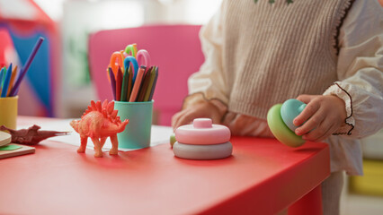 Little blonde girl stacks pastel toy rings with bare hands on a preschool building table amid colored pencils and a dinosaur toy  playful curiosity. © Krakenimages.com