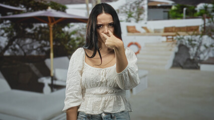 Fototapeta premium Young hispanic woman with crossed arms and frowning face near a resort pool building; frustration defiance.