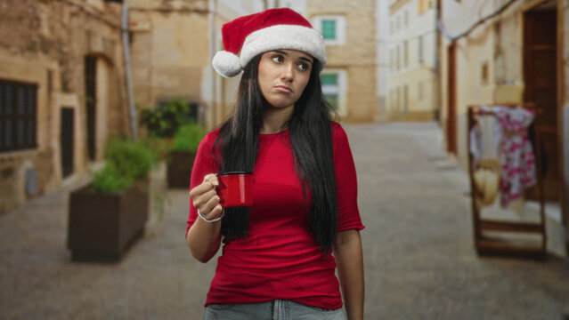 Woman in santa hat holds red mug with one hand, unsmiling and standing on a narrow street lined with shops and planters; tired solitude.