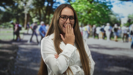 Obraz premium Woman with hand covering mouth laughing on a street, glasses and white shirt visible; playful amusement.
