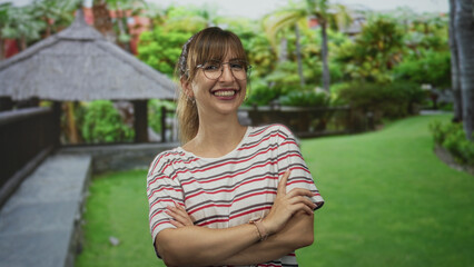 Naklejka premium Woman smiling with arms crossed beside thatched gazebo on forest pathway, wearing striped tee and glasses; confidence leisure summer.