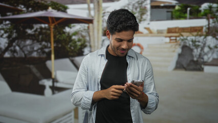 Fototapeta premium Man tapping smartphone with both hands and smiling while scrolling messages in studio set with pool backdrop and lounge chair visible; digital connection contentment.