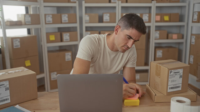 Man writing on sticky note beside parcel boxes and laptop in a delivery building; small business concentration.