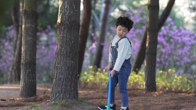 Little boy stands beside tree, holding tool. Soft sunlight filters through trees around him. Purple flowers bloom behind the trees in background. He appears focused, perhaps gardening or exploring