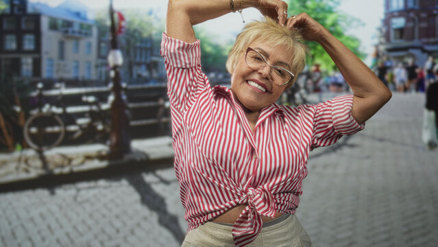 Senior hispanic woman forms heart with hands above head, tied red striped shirt and slight bare midriff on cobblestone street; joyful travel.