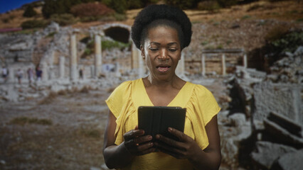 Obraz premium Woman holds tablet with both hands, tapping screen while standing among ancient ruins building and reading; curiosity travel discovery.