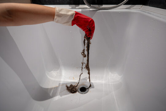 Person cleaning shower drain from trapped hair and debris to restore proper water flow. Hand with protective gloves removing hair in bathroom. Preventing clogs, improving drainage, keeping bath clean