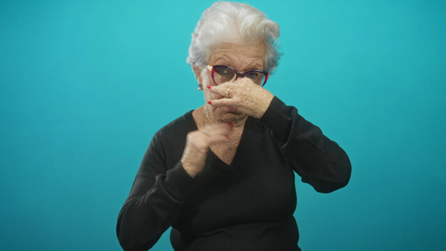 Elderly woman in black sweater pinches her nose with hand and closes eyes in turquoise studio beside glasses; disgust aversion.