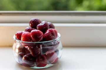 Fresh sweet cherries with water droplets in a glass jar on a white windowsill with green nature background © Dimitar
