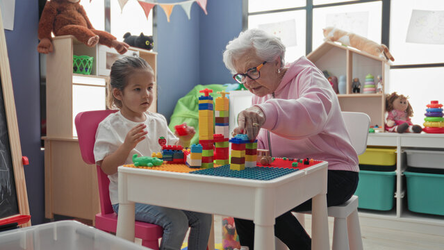 Senior woman and little girl build colorful block tower on a small table in a building playroom filled with toys; warmth and bonding.
