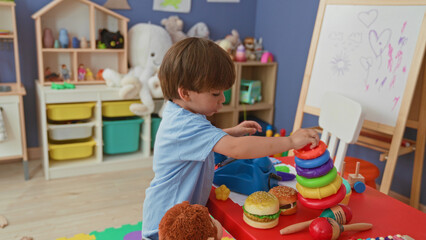 Fototapeta premium Boy barefoot holding and stacking colorful ring toy while standing on foam mat amid toy shelves and play kitchen in playroom building; playful joy.
