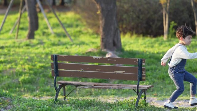 Little boy runs joyfully past a park bench. Sunlight dances across green grass and trees. Child's laughter echoes in peaceful park setting. Perfect for scenes of childhood innocence or leisurely walks