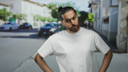 Fototapeta premium Man with hands on hips on street, tilted head and raised eyebrow in casual white tshirt, leaning forward and frowning; skepticism questioning.