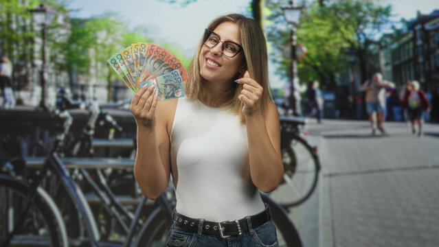 Woman holding fanned australian dollars on a street with bike racks, pedestrians and trees, wearing glasses and denim; playful wealth.