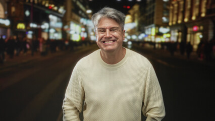Fototapeta premium Man smiling with hands in pockets on a busy city street at night, wool sweater and visible teeth, casual posture; contentment.