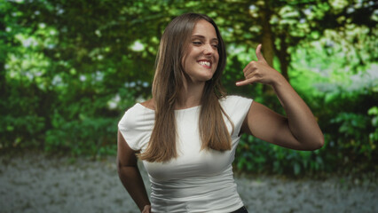 Fototapeta premium Woman making call me gesture with thumb and little finger extended, smiling in a forest; playful confidence.