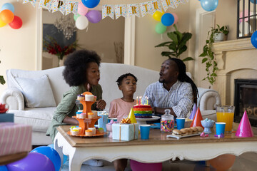 Obraz premium African American family blowing out birthday cake candles while sitting on sofa in living room