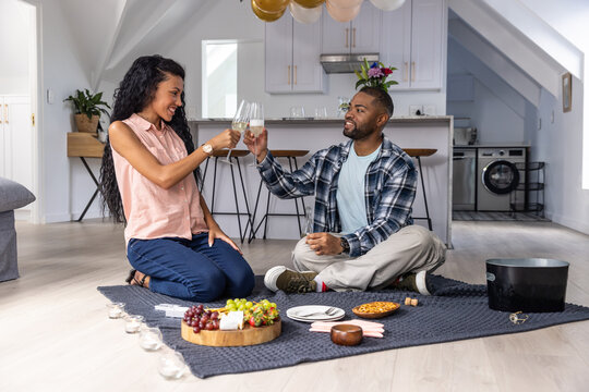 Couple sitting on knit picnic blanket in loft kitchen, toasting with stem glasses near charcuterie