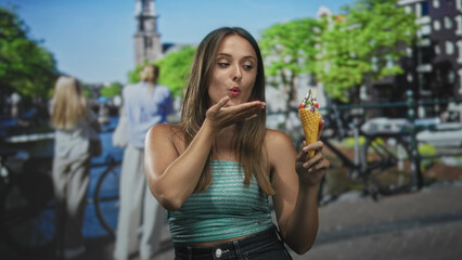 Fototapeta premium Woman holding ice cream cone on street, blowing toward cone and touching hair in casual pose; playful summer moment.