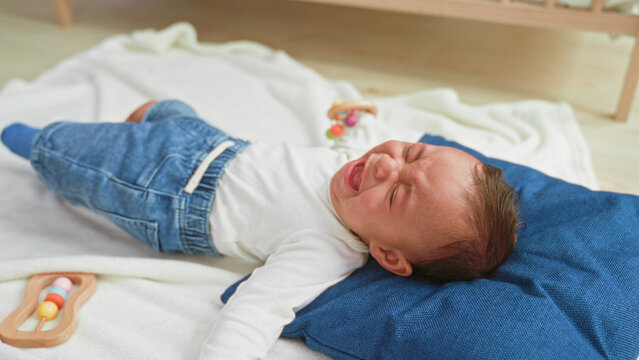 Toddler boy crying with tear and outstretched arm on blue pillow beside wooden rattle on blanket in building; distress seeking comfort.