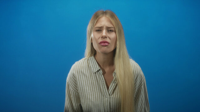 Woman with long blonde hair in striped shirt grimacing with puckered lips against blue studio backdrop; disgust discomfort.