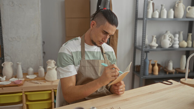 Man in apron writes in notebook holding pen and tapping chin while seated at pottery worktable with ceramic vases on shelves in studio; quiet contemplation.