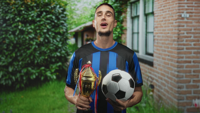 Man holding a soccer ball and gold trophy with bare hands while smiling in front of a brick building during an outdoor celebration; victory pride.