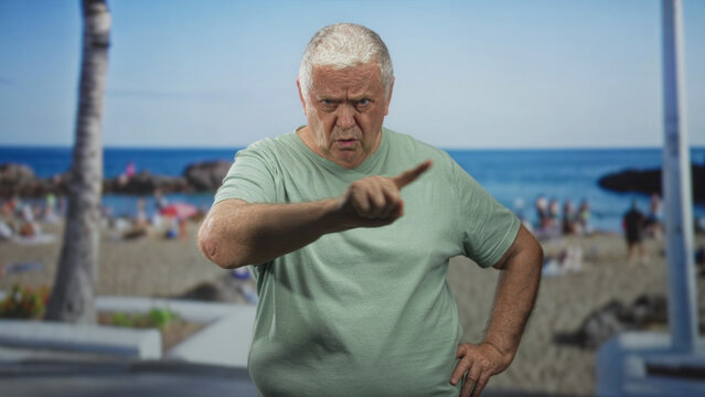 Senior man points finger to camera, hand on hip, standing on crowded beach promenade by palm tree; stern warning.