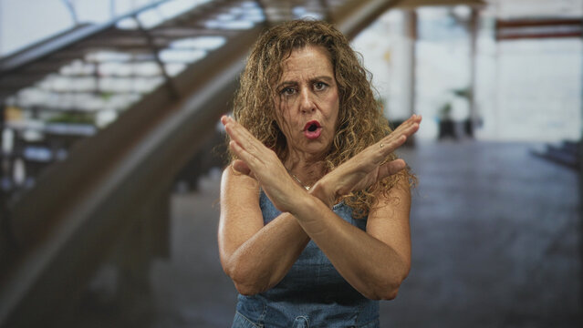 Middle aged hispanic woman with crossed forearms in an x gesture and raised index finger, frowning in an airport; defiance.